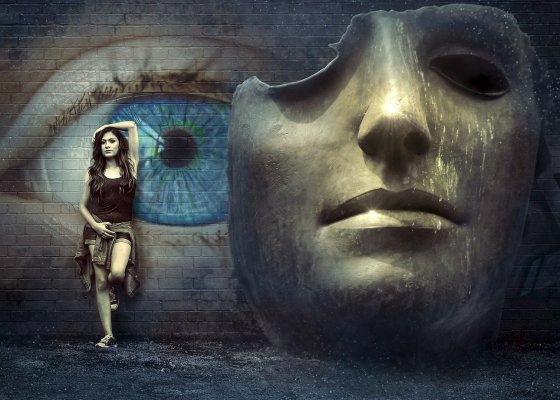 Description: Fantasy image. Large Surreal Mask in front of a blue eye painted on a wall. I girl is leaning on the wall to the left of the mask.