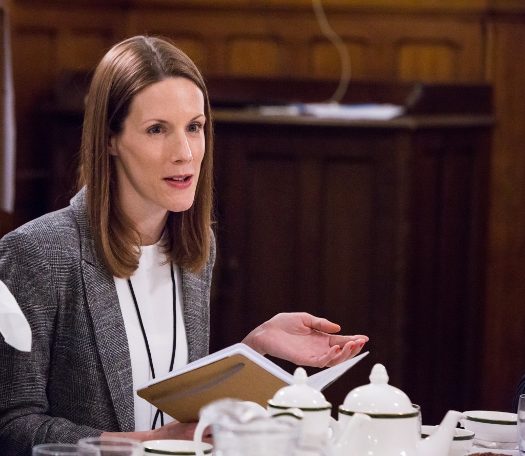 Me speaking at the event. I have shoulder-length brown straight hair and am holding an open notebook.