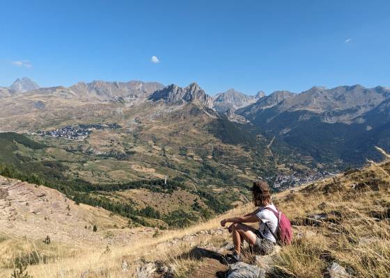 Picture of the back of a young woman sitting at the top of a mountain looking at a view of a valley surrounded by mountains.