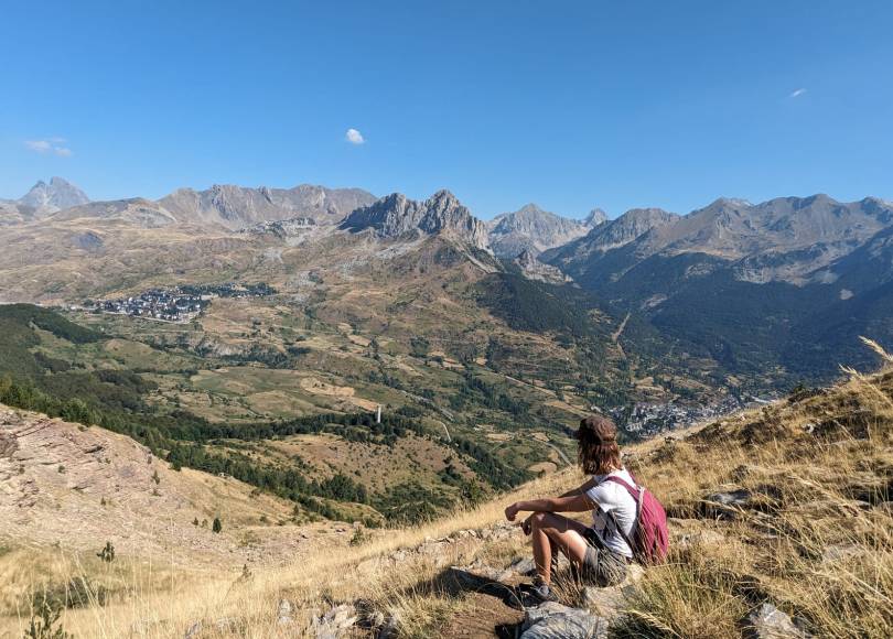 Picture of the back of a young woman sitting at the top of a mountain looking at a view of a valley surrounded by mountains.