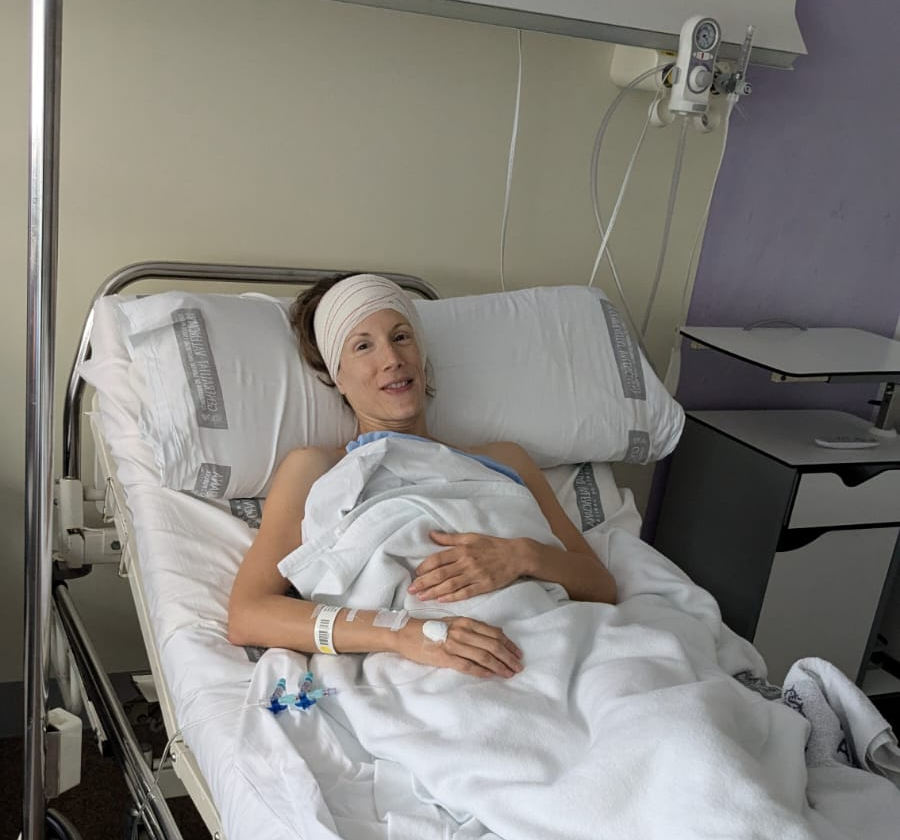 Photo of Carly post-surgery, sitting up slightly in bed with a bandage around her head smiling.