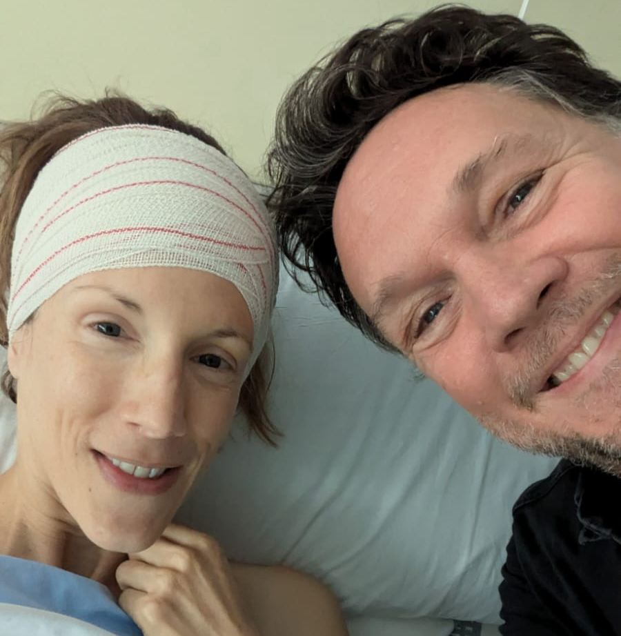 Photo of Carly in a hospital bed with a bandage around her head and her partner, both smiling.