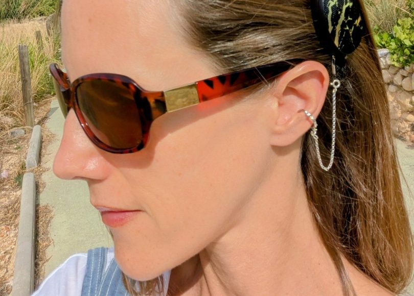 Close-up of the left side of Carly’s face and neck, outdoors on a country path. She has straight, shoulder-length brown hair and wears sunglasses. Her cochlear implant processor features Med-El’s black marble design with a gold pattern. The processor is secured to her ear by a Deafmetal hammered silver ring and chain.