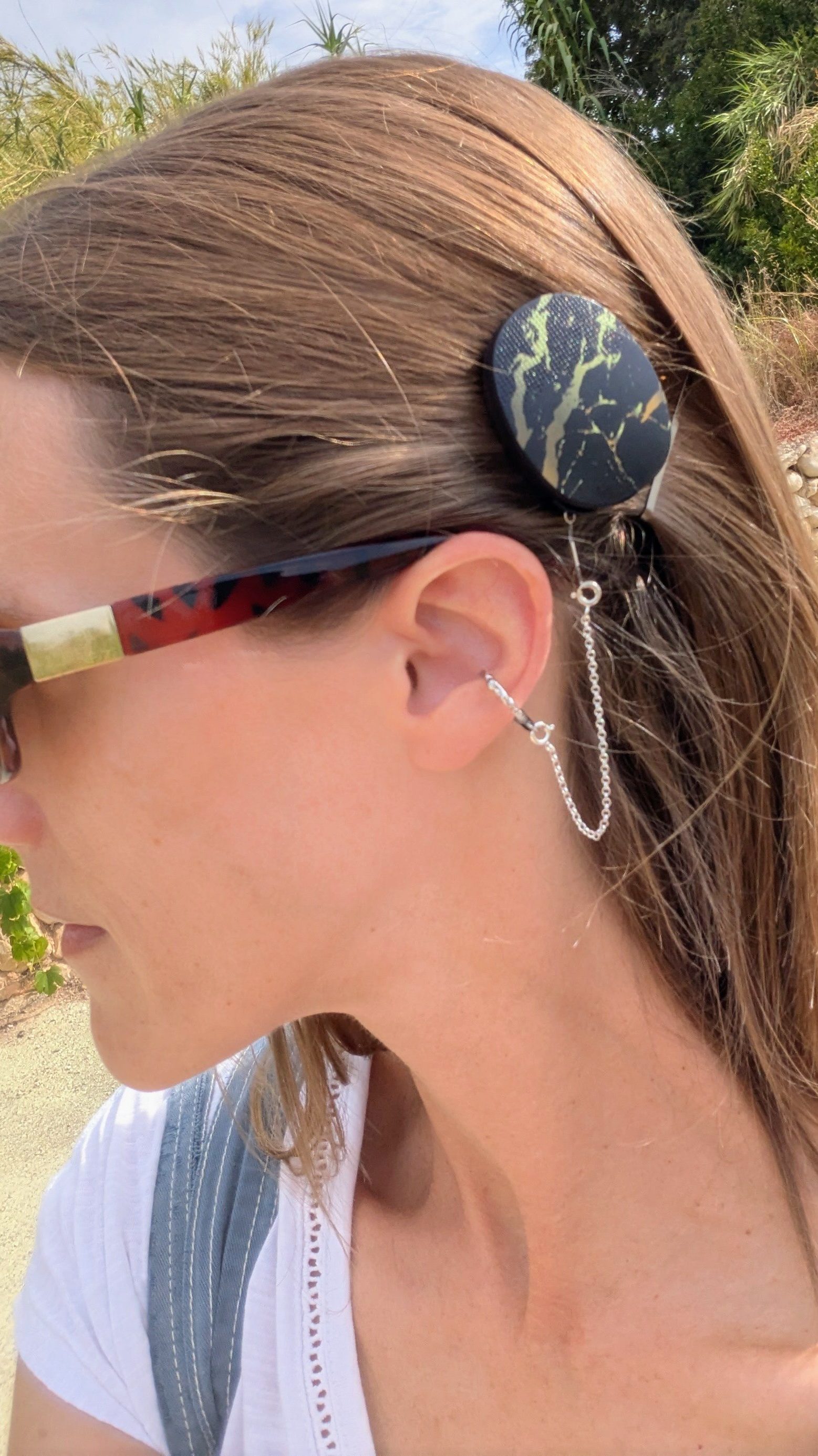Close-up of the left side of Carly’s face and neck, outdoors on a country path. She has straight, shoulder-length brown hair and wears sunglasses. Showing off her cochlear implant processor, featuring Med-El’s black marble design with a gold pattern, secured to her ear by a DeafMetal hammered silver ring and chain.