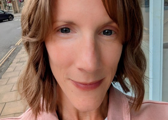 Carly, a woman in her early 40s, looking at the camera showing off her new wavy bobbed haircut. She is wearing a pink denim jacket and is outside with a street visible in the background.