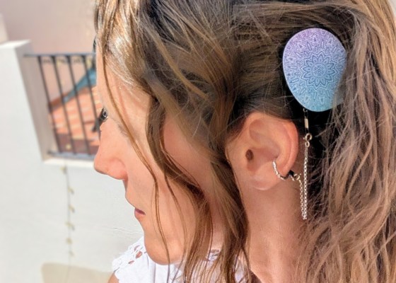 Profile of Carly. Her hair is wavy and she is wearing a blue and purple design cover on her Rondo 3 processor, and a silver safety chain connected to her ear with a ring. She is outside, looking over a terrace.