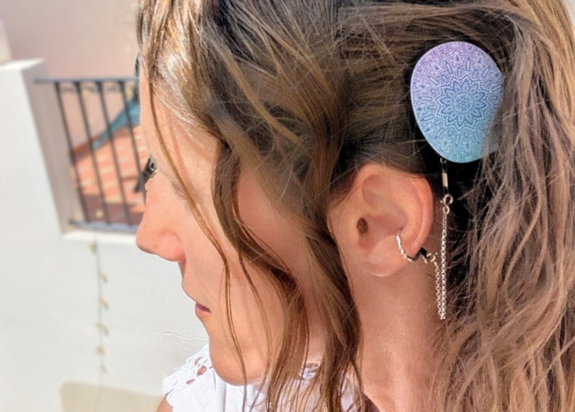Profile of Carly. Her hair is wavy and she is wearing a blue and purple design cover on her Rondo 3 processor, and a silver safety chain connected to her ear with a ring. She is outside, looking over a terrace.