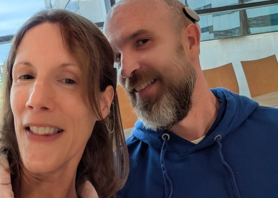 Carly and a guy smiling, showing off their Rondo 3 processors. Carly is wearing a pink jacket and the guy is mid-40s, with a grey beard, bald head, and a dark blue sweatshirt.