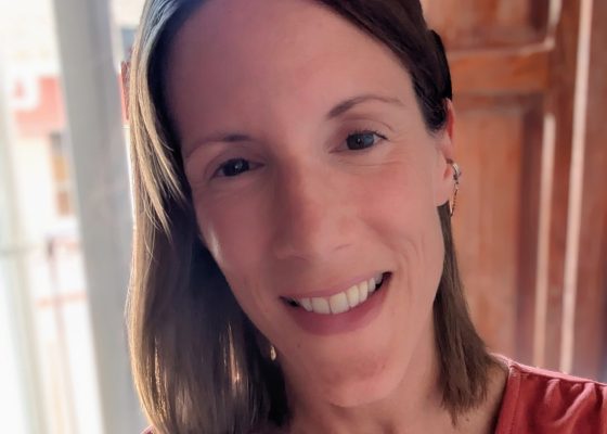 Photo of Carly in front of a window with Spanish-style wooden shutters. She is smiling, with her hair showing part of her cochlear implant processor. She is early 40s, with shoulder-length brown hair, and is wearing a dark coral coloured dress.